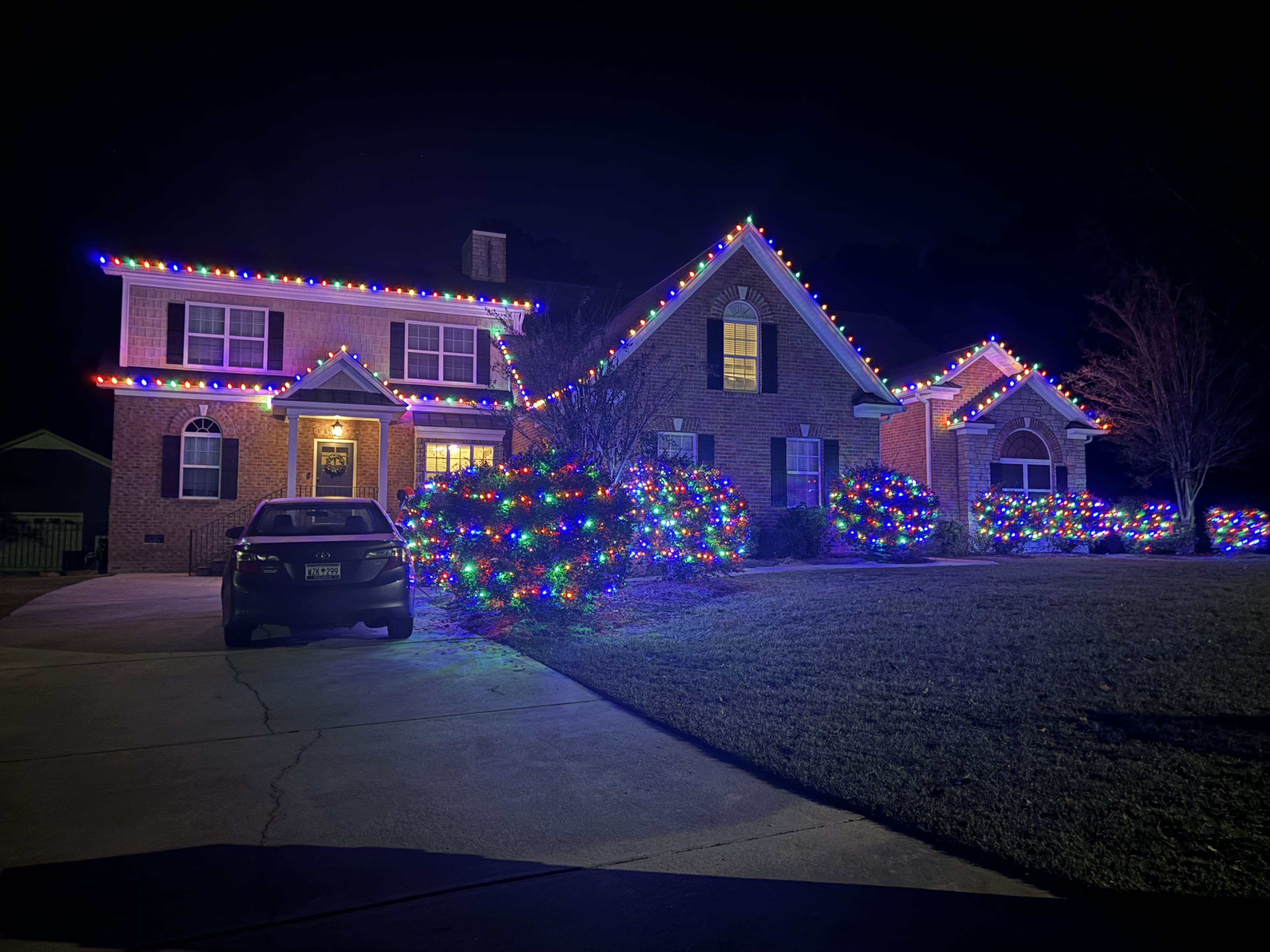 Festively decorated brick house with colorful lights, surrounded by illuminated shrubbery at night.